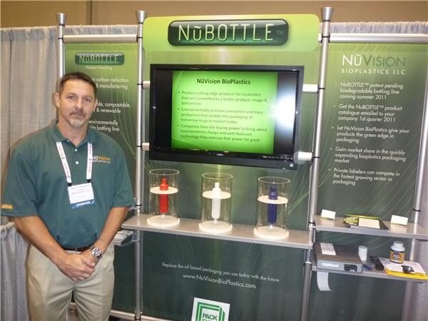 A man in a green polo with a lanyard stands in front of a NuBottle and NuVision BioPlastics LLC display with a screen and three bottles shown in glass domes.
