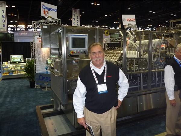 A mustached man in a dark vest and lanyard stands in front of a large stainless steel packaging machine, with Osgood and Bosch signs hanging overhead.
