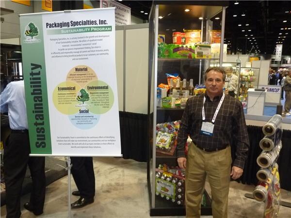 A man in a plaid shirt and lanyard stands between a Packaging Specialties, Inc. Sustainability banner and a shelving unit of packaged goods, with rolls of film at right.