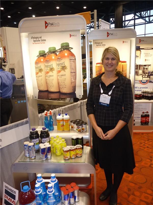 A smiling woman in a dark jacket and exhibitor badge stands by PakTech displays showing carrot juice bottles, Welch's bottles, Coca-Cola bottles and multipacks of cans and bottles.