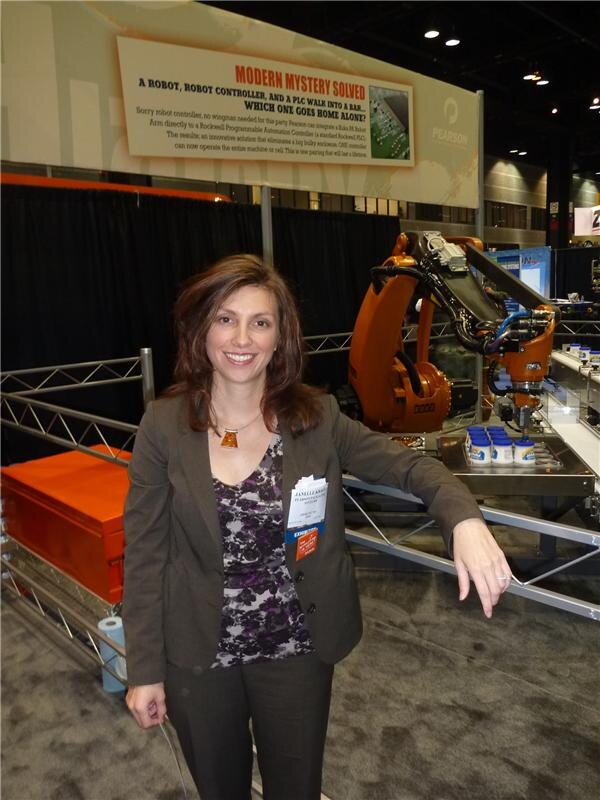A smiling woman in a gray jacket leans on a rail in front of an orange robotic arm and a Pearson banner reading Modern Mystery Solved, with small tubs on a conveyor.
