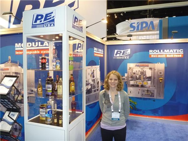A woman with curly hair and a lanyard stands in front of the P.E. Labelers USA booth with a glass case of bottles, Rollmatic signage, a labeling machine and a SIPA sign above.