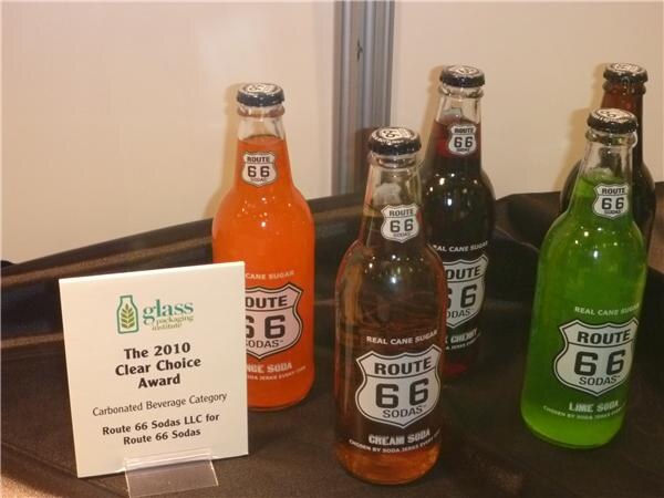 Five glass bottles of Route 66 Sodas in orange, cream soda, cherry and lime on a dark tablecloth, beside a 2010 Clear Choice Award sign for the carbonated beverage category.