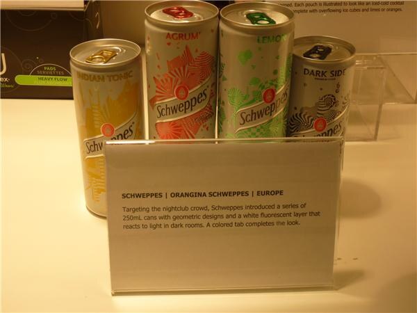 Four slim Schweppes cans with geometric designs, labeled Indian Tonic, Agrum, Lemon and Dark Side, behind a sign about Orangina Schweppes in Europe.