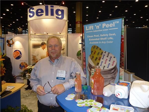 A man in a gray shirt holds his glasses at a blue table with bottles, jugs and seal samples, beside a Selig Lift 'n' Peel induction liner banner.
