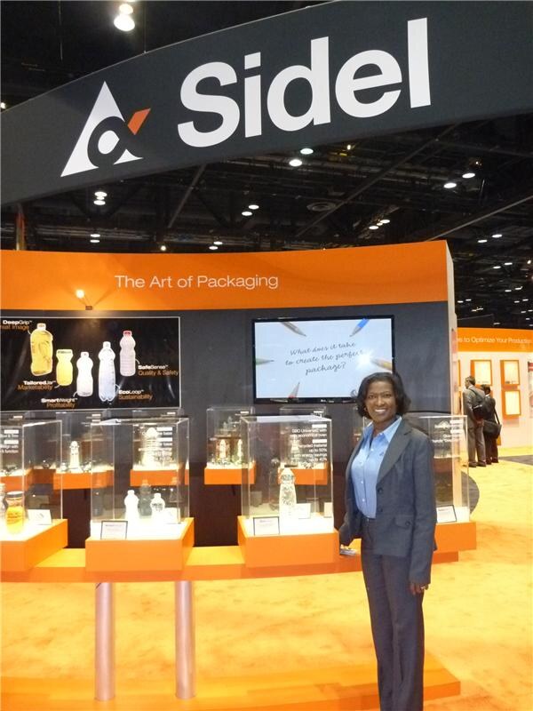 A woman in a gray blazer and blue shirt stands in front of the orange Sidel booth, with bottle displays in glass cases and a screen reading "The Art of Packaging" behind her.