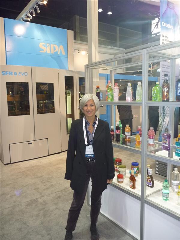 A woman with short gray hair and a lanyard stands between a SIPA SFR 6 EVO machine and a shelf of bottles, including Pepsi bottles, in the SIPA booth.
