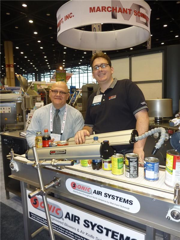 Two men stand at a Sonic Air Systems conveyor display holding canned goods and bottles, under a Marchant Schmidt, Inc. sign. One wears a lanyard and tie, the other a dark polo.