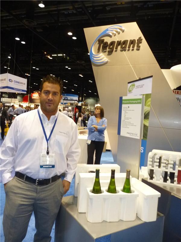 A man in a white shirt with an exhibitor badge stands by foam wine bottle shippers at the Tegrant booth, with Protexic signage and a ConeDrive banner in the background.