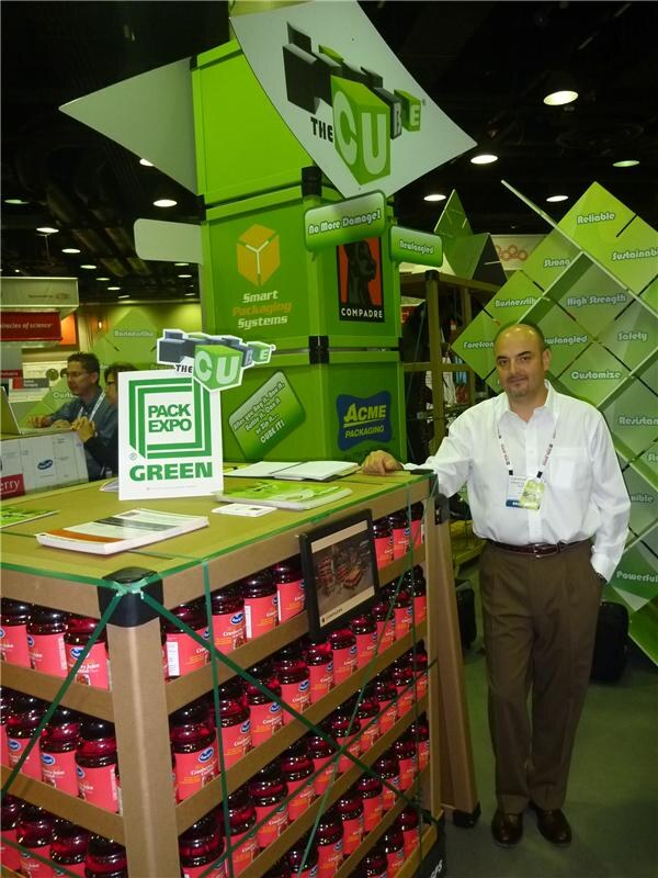 A man in a white shirt stands beside a pallet display of Ocean Spray cranberry jars wrapped in green strapping, under green The Cube signs with Compadre and Acme Packaging logos.