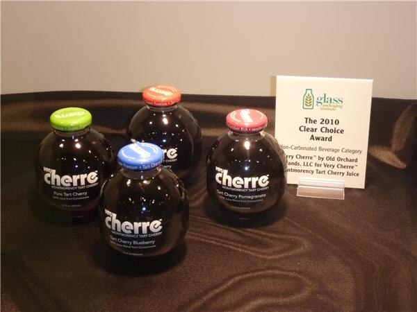 Four round dark bottles labeled Cherre with green, blue, orange and red caps sit on dark fabric beside a Glass Packaging Institute 2010 Clear Choice Award sign.