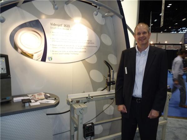 A smiling man in a dark blazer and light striped shirt stands in the Videojet booth beside a conveyor and printer, with a Videojet 1610 display lit on the wall behind him.