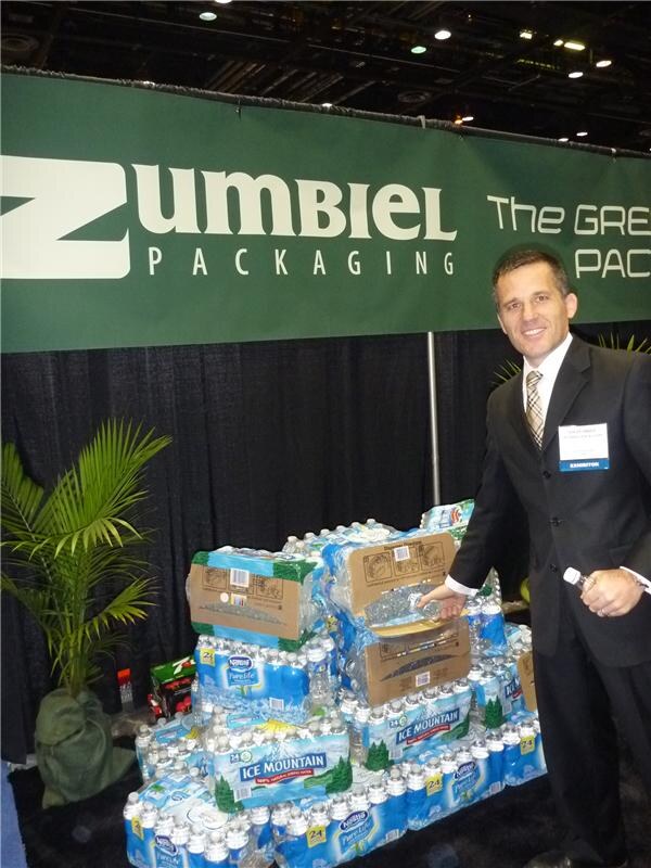 A man in a dark suit with an exhibitor badge stands beside a stack of Ice Mountain and Nestlé Pure Life bottled water packs under a green Zumbiel Packaging banner.