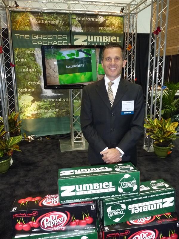 A man in a dark suit with an exhibitor badge stands behind green Zumbiel Packaging Tree Hugger cartons holding Dr Pepper Cherry cans, in front of a screen reading Sustainability.