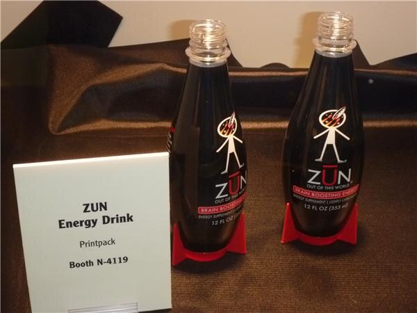 Two dark ZUN brain boosting energy drink bottles with open necks stand on red bases on brown fabric, beside a sign reading ZUN Energy Drink, Printpack, Booth N-4119.