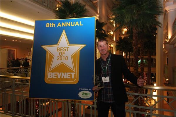 A man in a dark jacket and plaid shirt holds an award and gives a thumbs up beside a blue and gold 8th Annual BevNET Best of 2010 sign sponsored by Wild, in a hotel atrium with palm trees.