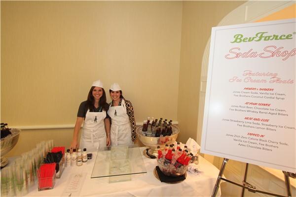 Two women in white aprons and paper caps stand behind a table of bottles and glasses at the BevForce Soda Shop, beside a menu sign listing ice cream soda floats.