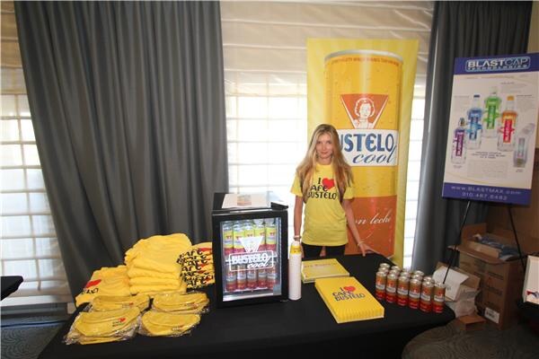 A woman in a yellow Café Bustelo T-shirt stands behind a table with folded shirts, a small cooler and canned drinks, beside a Bustelo cool banner and a BlastCap poster.