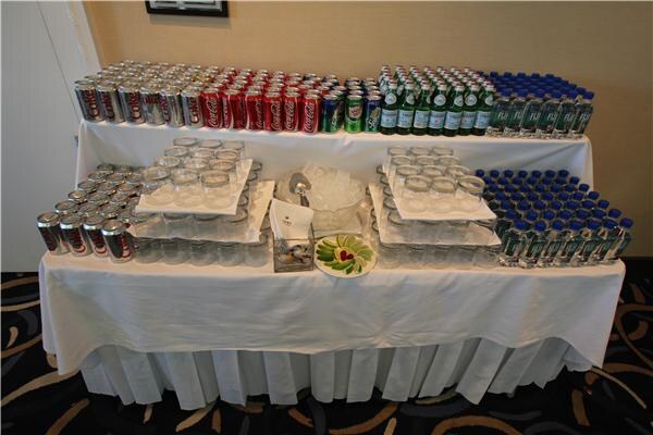 A tiered table with white cloth holds rows of Coca-Cola and other soda cans, green glass bottles, bottled water, stacks of clear glasses, an ice bowl and a small garnish plate.