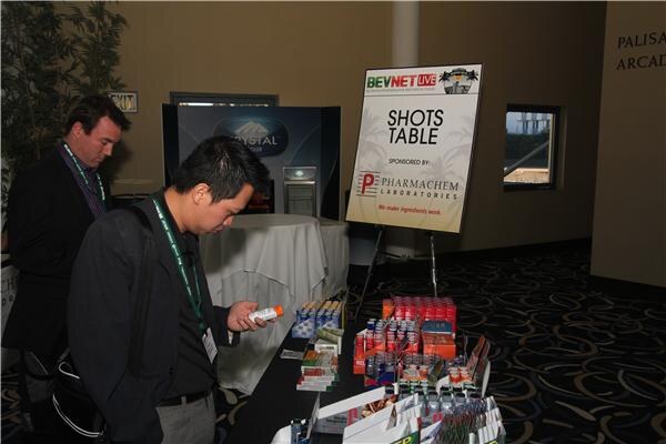 A man in a dark jacket looks at a small bottle beside a table of shot products under a Shots Table sign sponsored by Pharmachem Laboratories. Another man stands behind him.