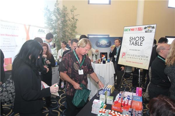 Attendees stand around a table of shot products beside a Shots Table sign sponsored by Pharmachem Laboratories. A Crystal Cooler display is in the background, and a man holds a green bag.