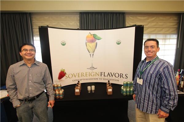 Two smiling men stand on either side of a Sovereign Flavors booth with a backdrop showing a glass and fruit, with small bottles and cans arranged on a black table.