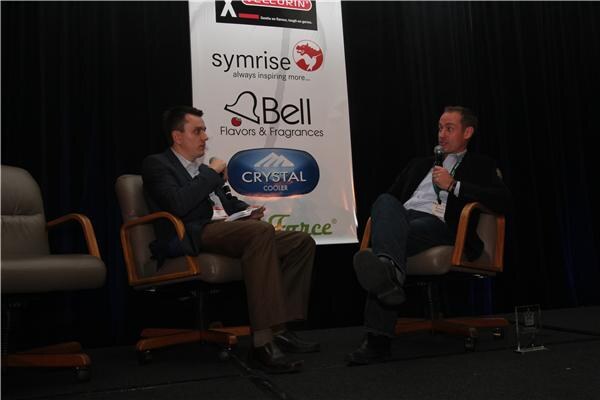 Two men holding microphones sit in armchairs on a dark stage in front of a banner with Velcorin, Symrise, Bell Flavors & Fragrances and Crystal Cooler logos.