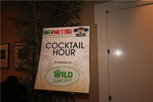 A sign on an easel reads BevNET Live Cocktail Hour, sponsored by Wild, with the Wild logo, placed near a plant and a door.