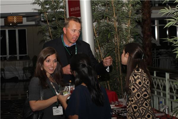 A man in a dark blazer with a lanyard gestures while talking with two women in the foreground and one at right, at a cocktail reception with bamboo plants and a Moss-Adams banner behind them.