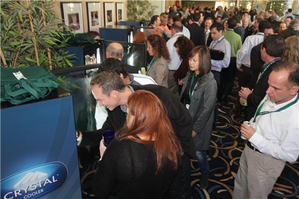 A crowd of lanyard-wearing attendees gathers around a sample bar with glass-front coolers, in the foreground a blue Crystal Cooler unit, as people lean in to look and hold cups.