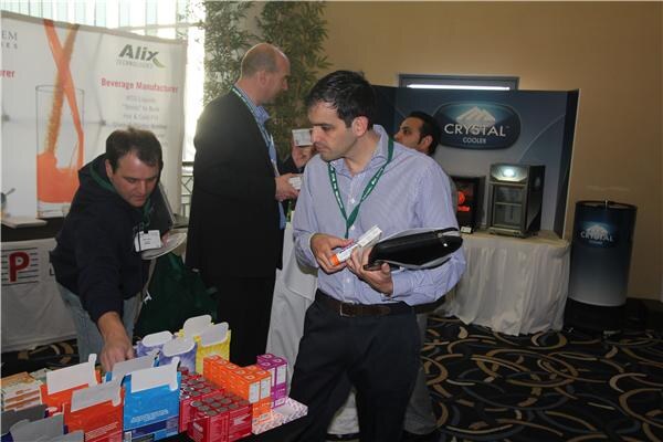 Men in lanyards stand at a table of boxed shot products in front of an Alix Technologies banner and a Crystal Cooler display; one man bends to pick up a box and another holds a package.