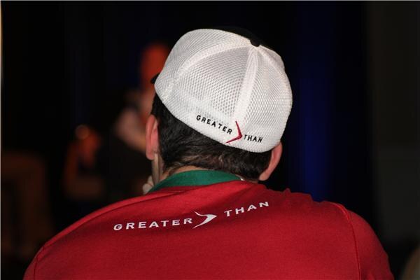 View from behind of a man in a white Greater Than cap and red Greater Than shirt, with a green lanyard, facing a dark, blurred stage area.