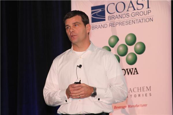 A man in a white dress shirt with a lapel microphone speaks and holds a clicker in front of a banner reading Coast Brands Group, Brand Representation.
