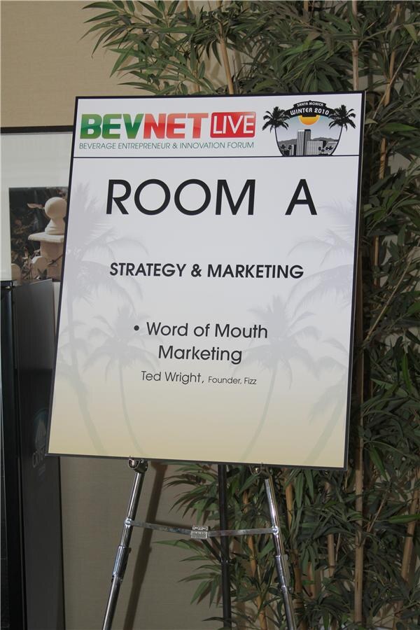 Easel sign for Room A, Strategy & Marketing, listing the session Word of Mouth Marketing with a speaker from Fizz, set in front of bamboo plants.