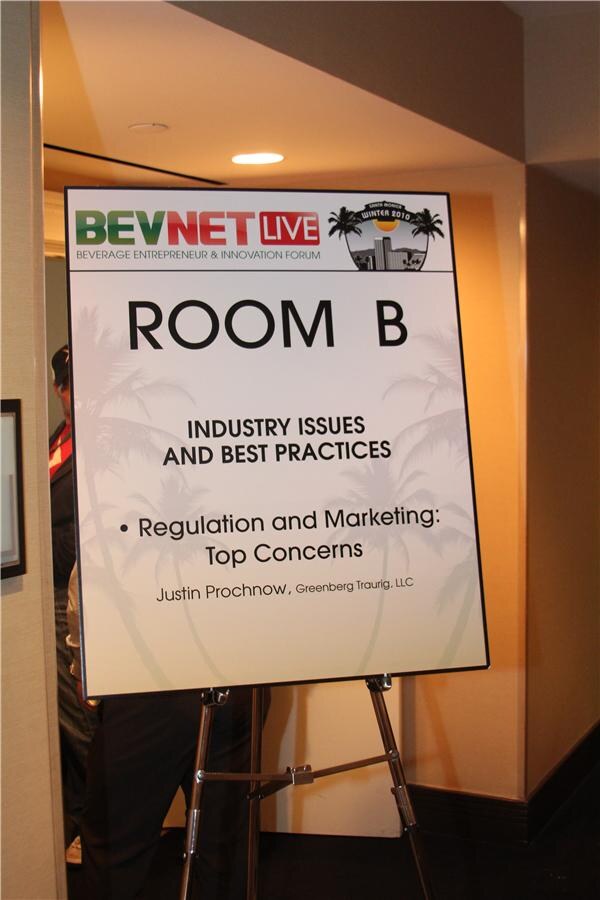 Easel sign for Room B, Industry Issues and Best Practices, listing the session Regulation and Marketing: Top Concerns with a speaker from Greenberg Traurig, LLC, in a hotel hallway.