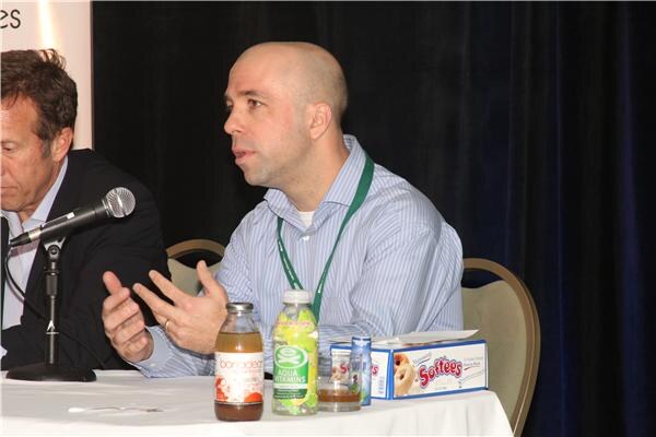 A bald man in a striped shirt and green lanyard gestures while speaking at a panel table, with Aqua Vitamins and other bottles and a Softees box in front of him; another man is partly visible at left.