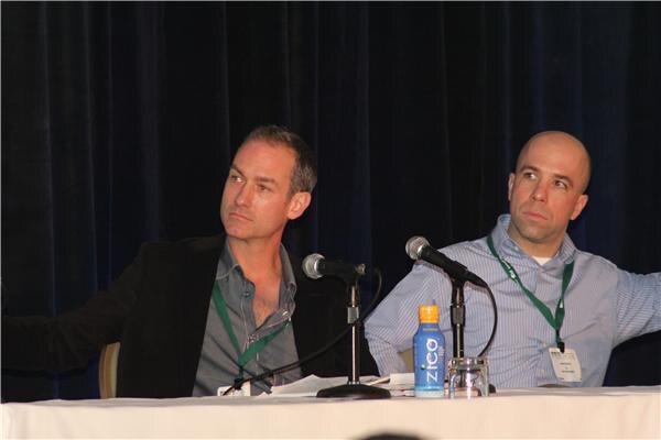 Two men with green lanyards sit at a draped table with microphones and look off to the side; a bottle of Zico coconut water sits between them.