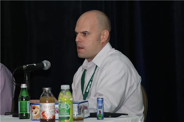 A man in a white shirt and green lanyard sits at a panel table beside a microphone, with several beverage bottles including Aqua Vitamins and a box of snacks in front of him.