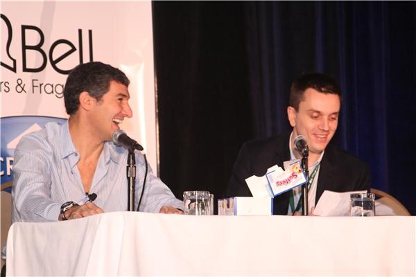 Two men sit at a draped table with microphones on a stage. The man on the left laughs; the man on the right looks down at papers. A Softees tissue box sits on the table and a Bell banner is behind.