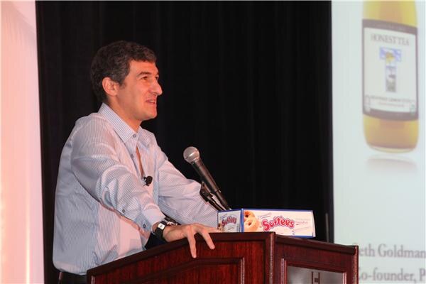 A man in a light blue striped shirt speaks at a wooden podium with a microphone. A Softees box sits on the podium, and a projected slide beside him shows an Honest Tea bottle.
