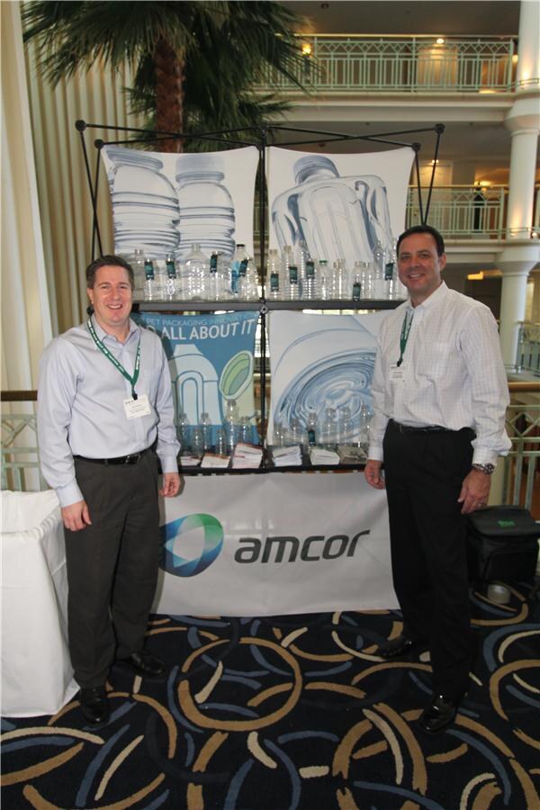 Two men in dress shirts and lanyards stand on either side of an Amcor display of clear PET bottles and packaging posters, with an Amcor logo banner, in a hotel atrium.