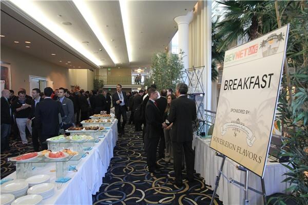 Attendees in business attire stand and mingle along buffet tables in a hall, next to an easel sign reading Breakfast, sponsored by Sovereign Flavors.