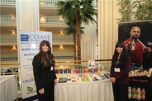 Two women with lanyards stand beside a draped table of beverages in front of a Coast Brands Group banner listing Celsius and New Leaf, with a poster of a man holding a bottle at right.