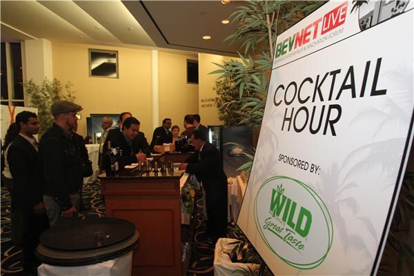 A large BevNET Live Cocktail Hour sign sponsored by Wild stands beside a bar where several people in dark clothing gather and a bartender stands behind the counter.