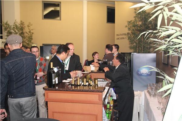 People stand and talk around a wooden bar with wine bottles while a bartender serves; a Crystal Cooler display stands against the wall and glasses sit on a table at right.