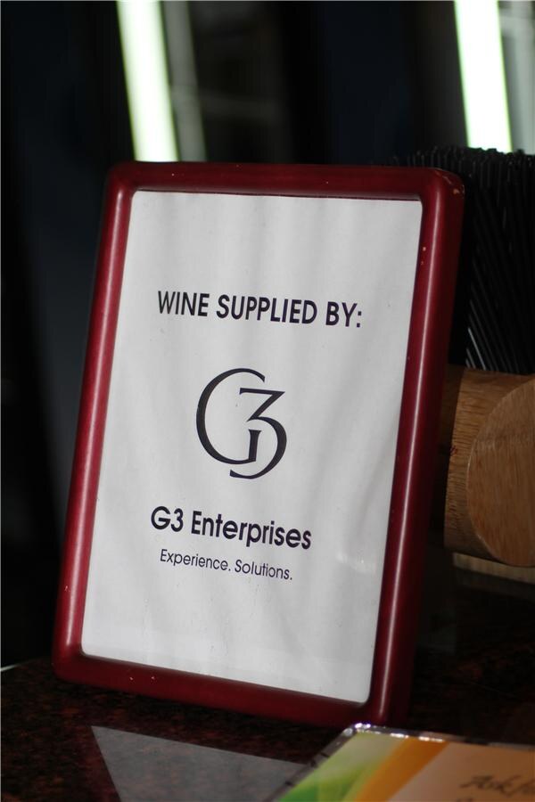A framed sign in a red holder reads "Wine supplied by: G3 Enterprises, Experience. Solutions." with the G3 logo, placed on a bar counter.
