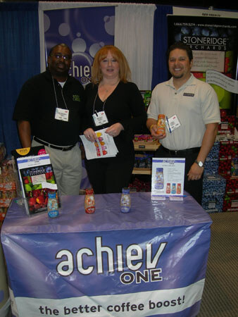 Two men and a woman with name badges pose behind a table with an achiev ONE banner reading 'the better coffee boost!'. Bottles sit on the table and a Stoneridge Orchards sign hangs behind.