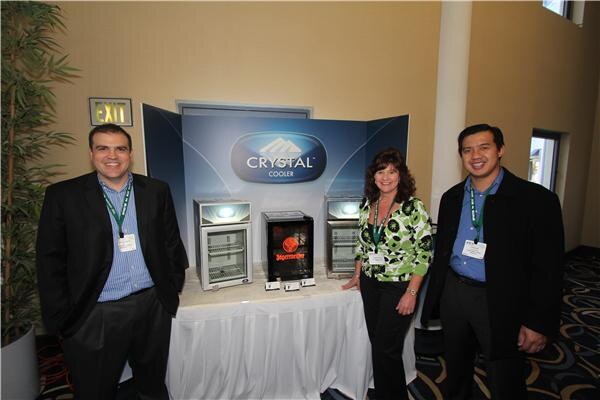 Two men and a woman with lanyards stand beside a table holding small Crystal Cooler refrigerators, one branded Jägermeister, in front of a blue Crystal Cooler backdrop.