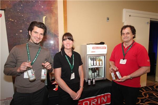Two men and a woman with lanyards stand at a table with a Velcorin-branded mini refrigerator of bottled drinks; the men each hold two small bottles.