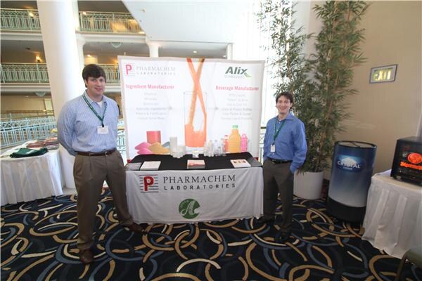 Two men with lanyards stand at a Pharmachem Laboratories table under an Alix banner showing ingredient and beverage manufacturing, with a Crystal Cooler unit at right.